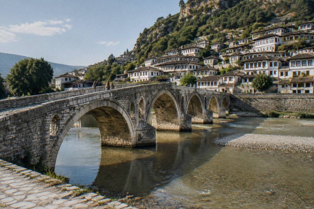 Stone arches of the Gorica Bridge crossing the Osum River toward old houses on the far bank