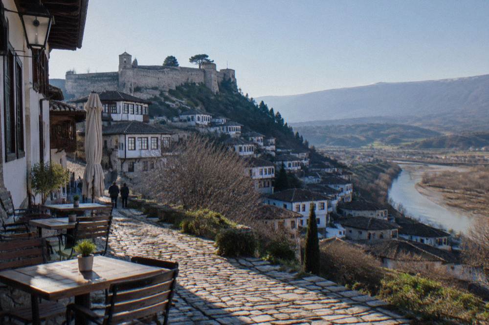 Outdoor restaurant tables on a cobbled Berat street with white Ottoman houses rising behind