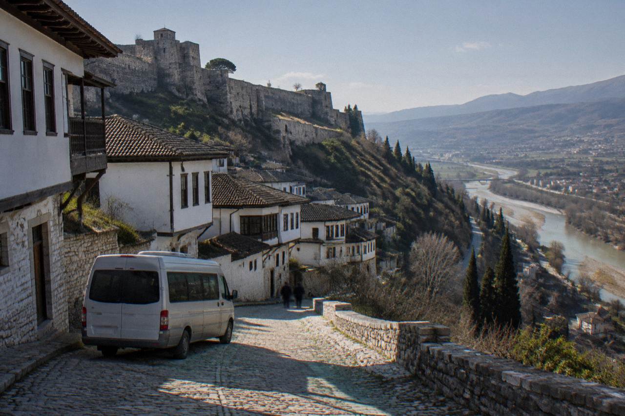 A small minibus parked on a stone street below the whitewashed houses of an Albanian hill town