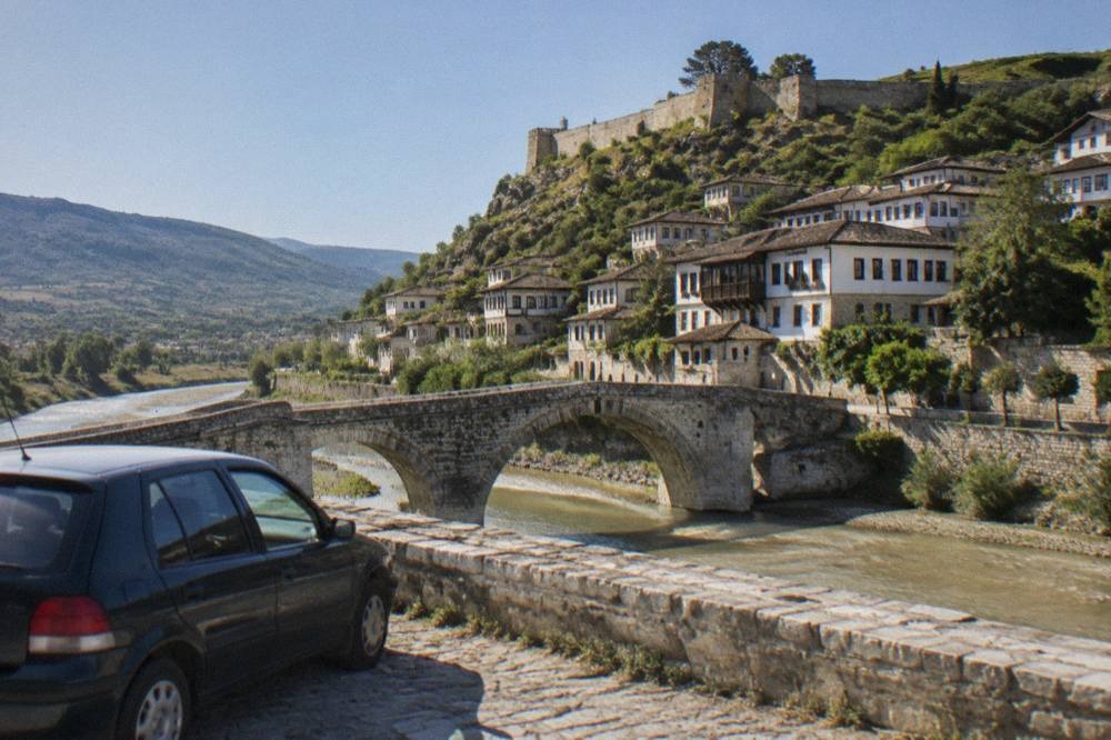 A parked car beside a stone bridge over the Osum River in Berat with Ottoman houses on the hillside above