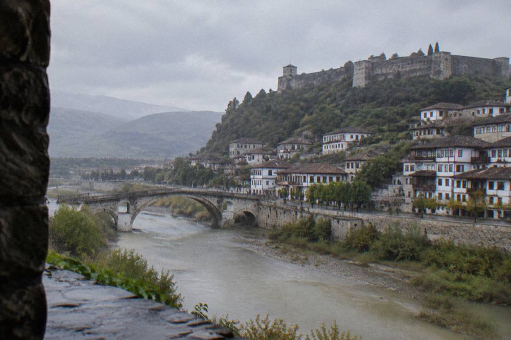 A stone bridge over a river with a walled hilltop fortress above a town