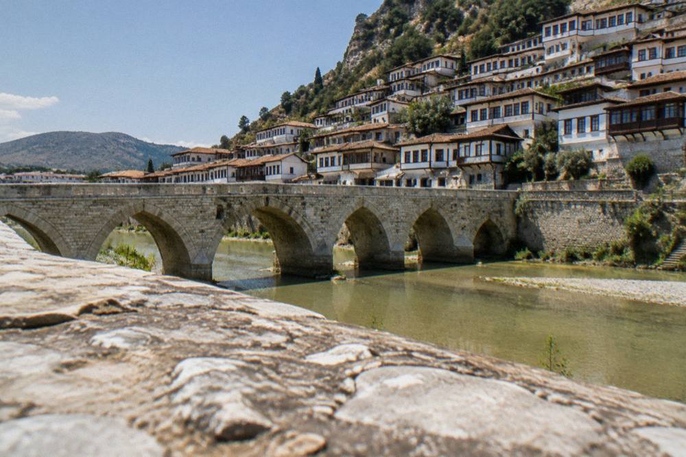 A stone arch bridge with many pointed arches crossing a green river between old houses