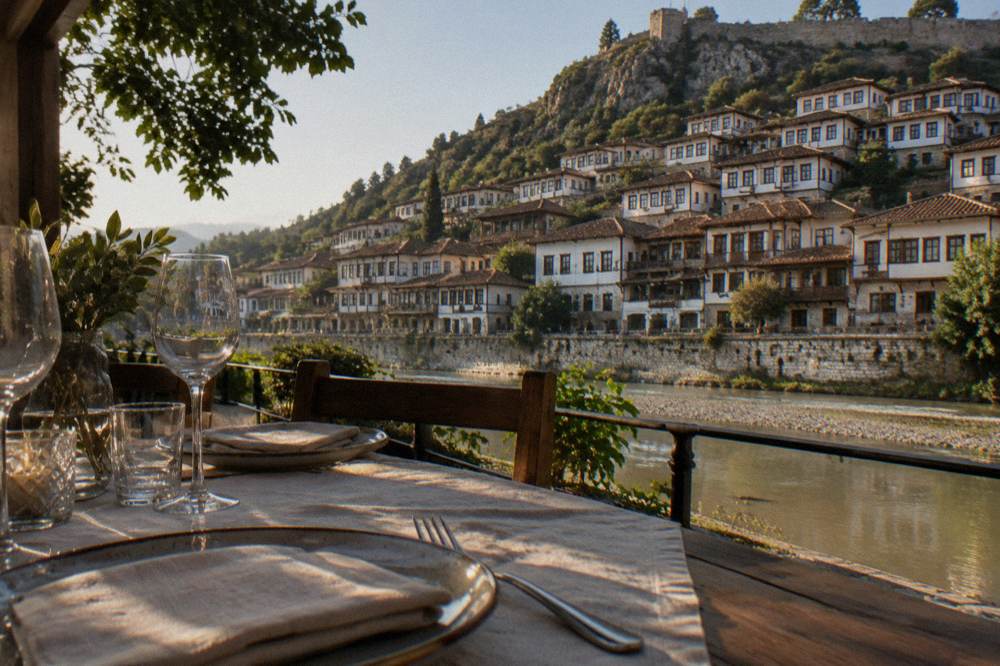 A riverside terrace table set for dinner with old houses across the water