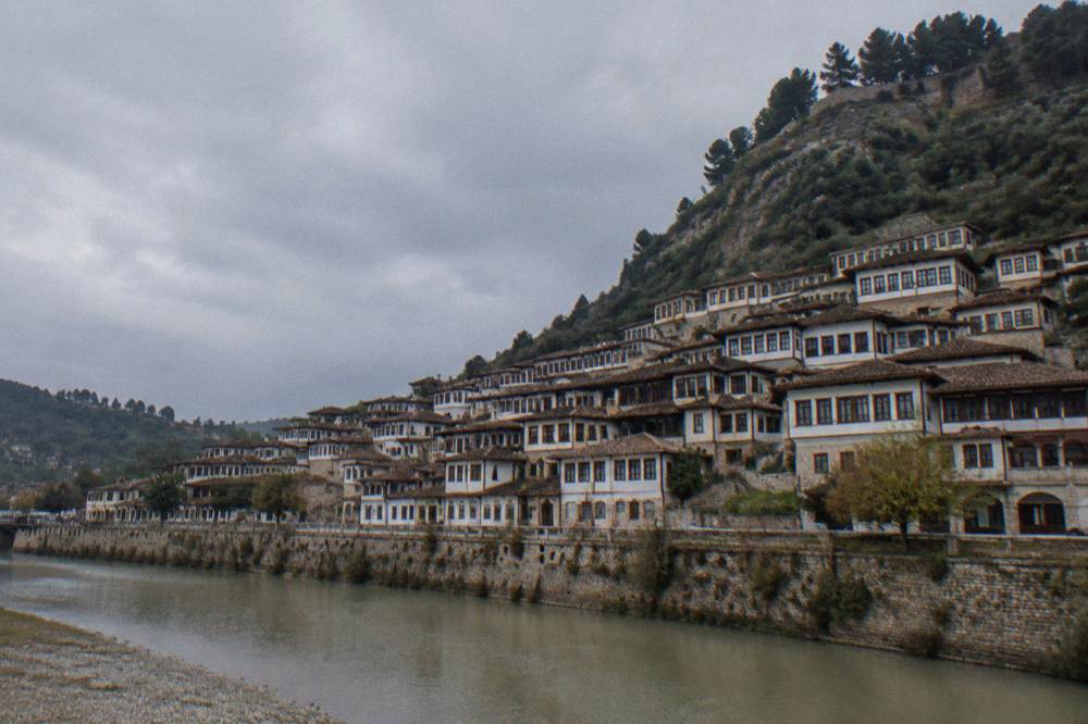 Whitewashed Ottoman houses with many windows stacked on a green hillside above a calm river