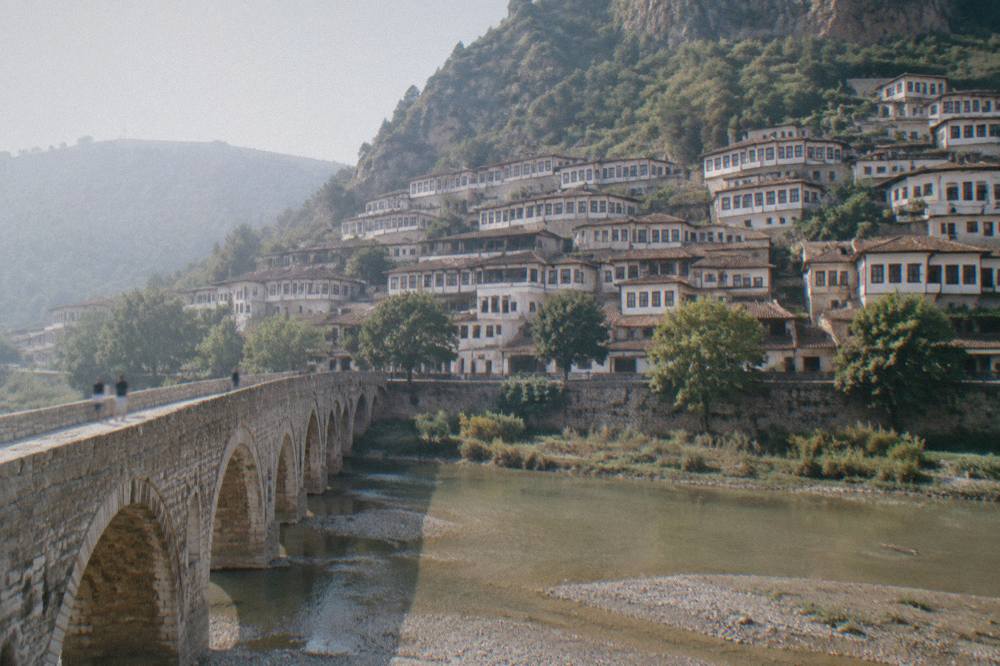 View across the Osum River from Gorica Bridge showing the stacked windowed houses of Mangalem on the hillside