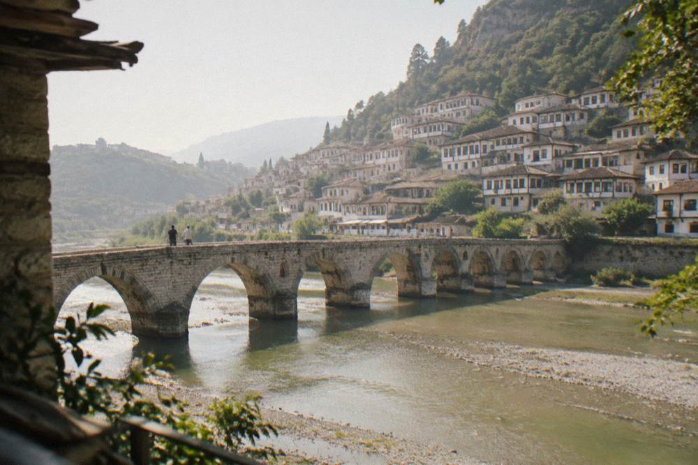 Old stone bridge with arched spans crossing a green river between two hillside towns of white Ottoman houses