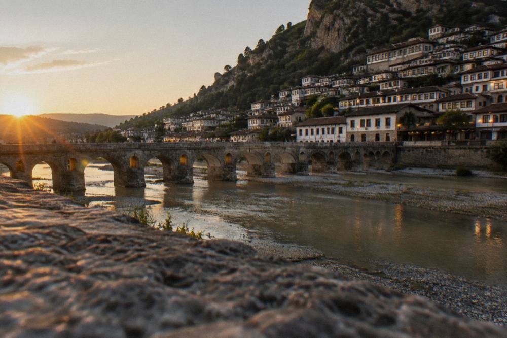 Wide view of a river town at dusk with a stone bridge lit softly and lights appearing in windows on both banks