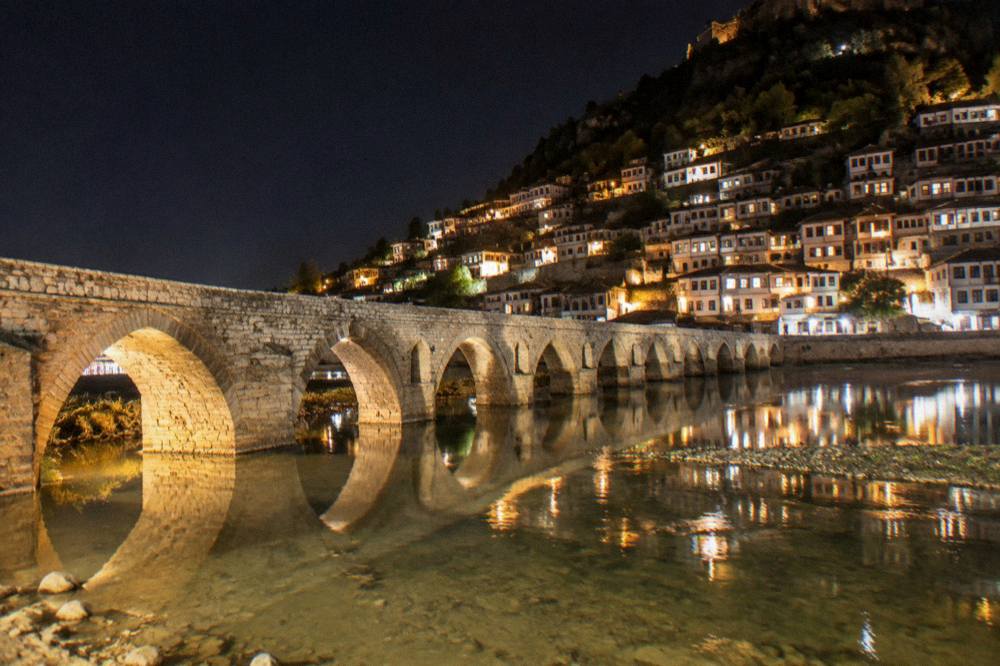 Night view of a stone bridge and the lit windows of a hillside town reflected in a calm river