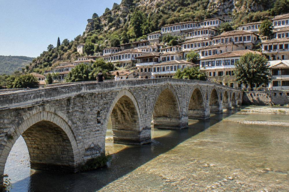 The stone arches of the Gorica Bridge over the Osum River with Mangalem's hillside houses behind
