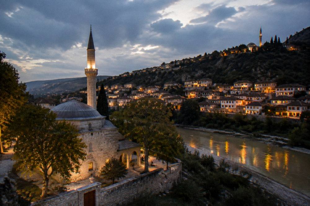 Night view of Berat's hillside with lit windows and a mosque silhouette above the dark river