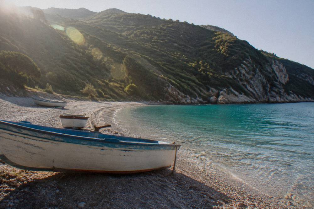 A pebble cove with clear turquoise Ionian water and green hills rising behind the shore
