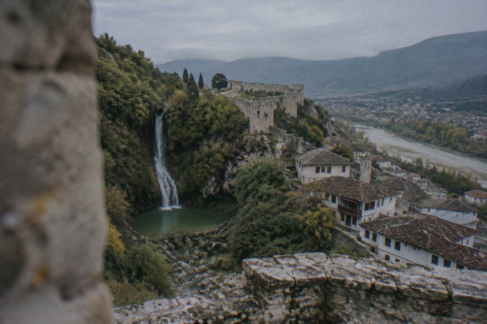 A narrow waterfall dropping down a wooded rock face into a small green pool in the hills above Berat