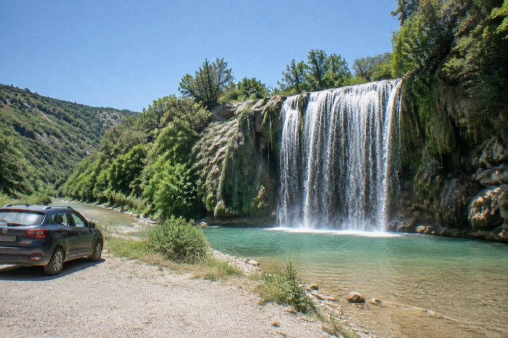 A parked car on a gravel pull-off beside a forested valley road in Albania