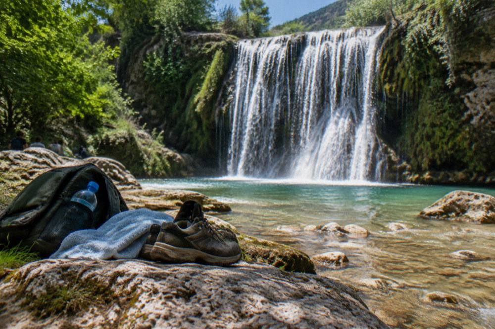 A small daypack with a water bottle, towel and walking shoes set on a stone beside a stream