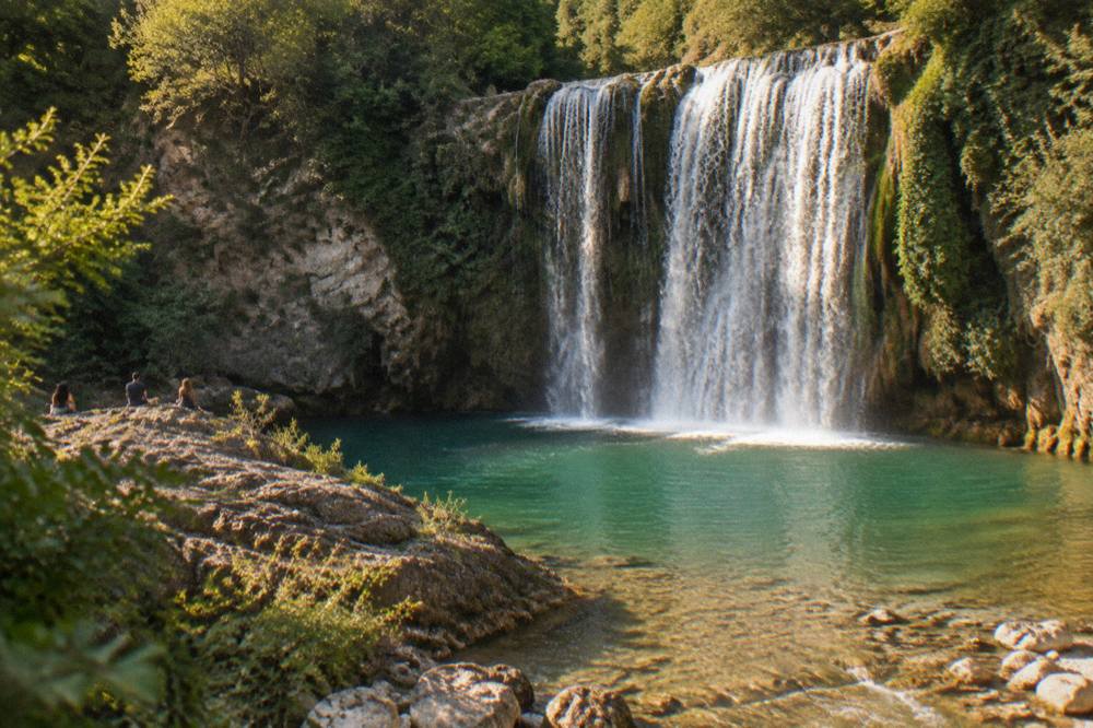 A clear green pool at the base of a waterfall surrounded by smooth wet rock and trees