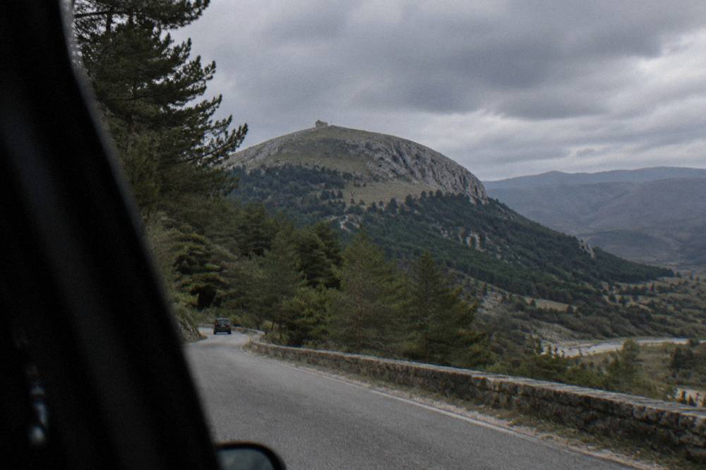 A narrow asphalt mountain road climbing through pine forest toward the Tomorr highlands