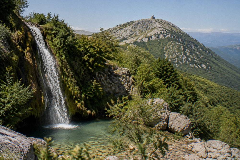 A waterfall dropping over a mossy rock face into a clear forest pool on the flank of Mount Tomorr