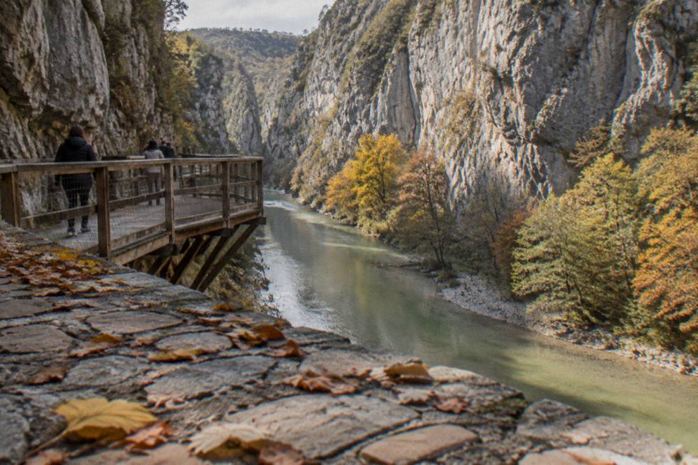 Steep limestone canyon walls above a calm green river, with autumn trees along the waterline at Osumi Canyon.