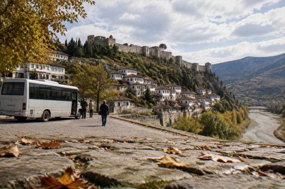 A small regional bus parked at a roadside terminal in Albania with hills behind it in autumn.