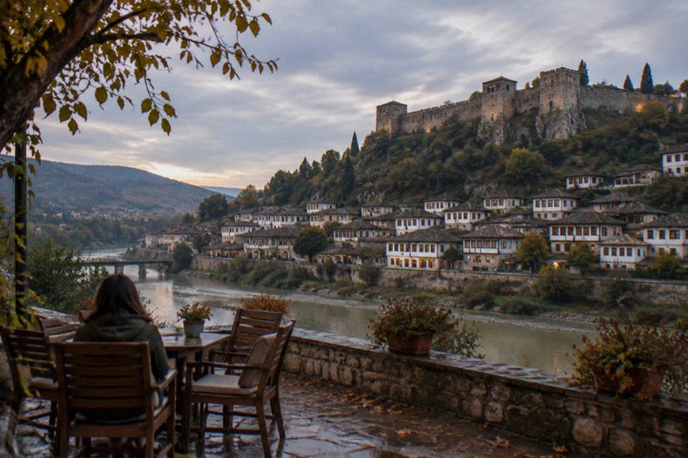 Guesthouse terrace with wooden chairs overlooking the lit windows of old houses across the river in Berat.