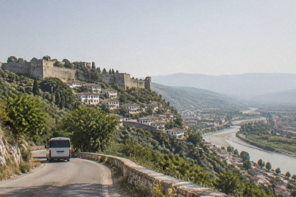 A minibus travelling along a winding road through green hills toward a river valley town
