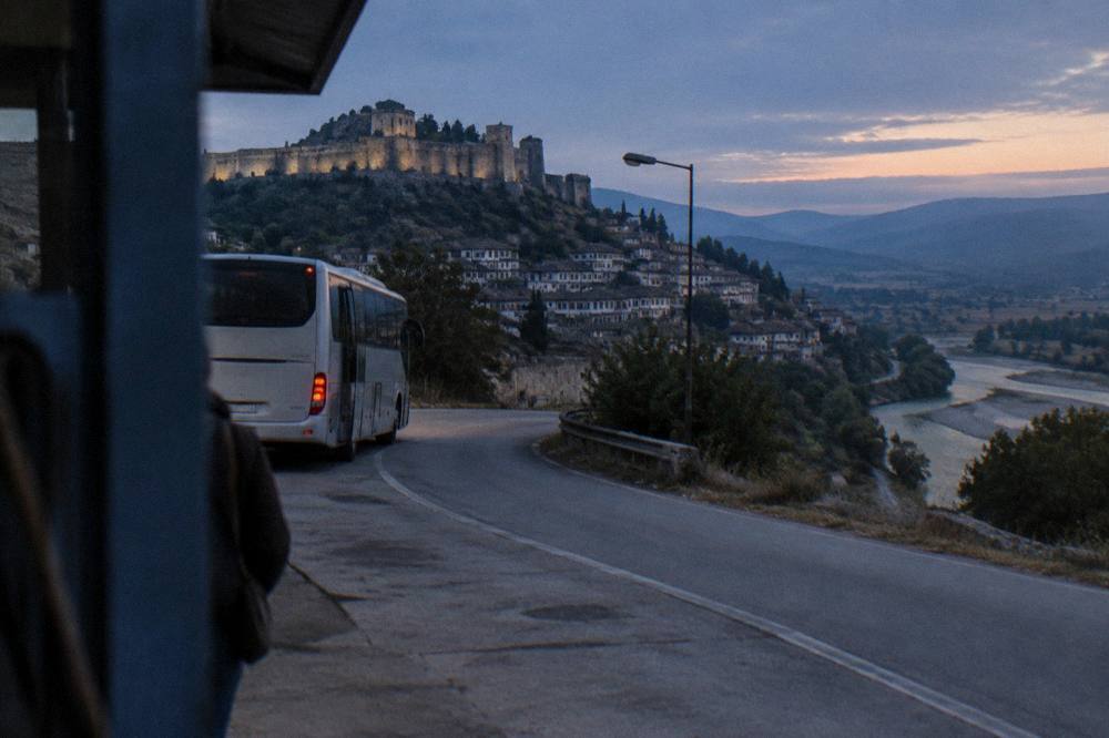 A bus departing a roadside terminal in the early morning with the road curving away between dry hills
