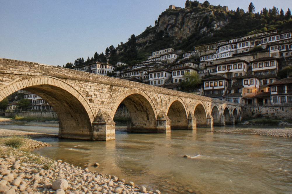 Berat's Gorica Bridge over the Osum River at dusk with lit windows above the water