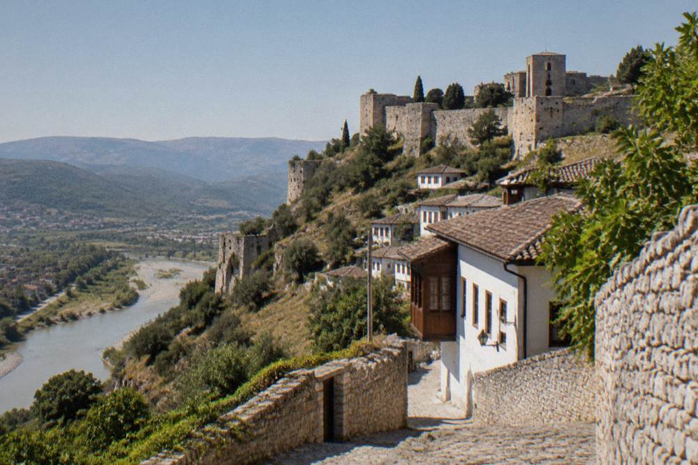Stone walls and towers of Berat Castle seen from a lane below with the hillside dropping toward the river