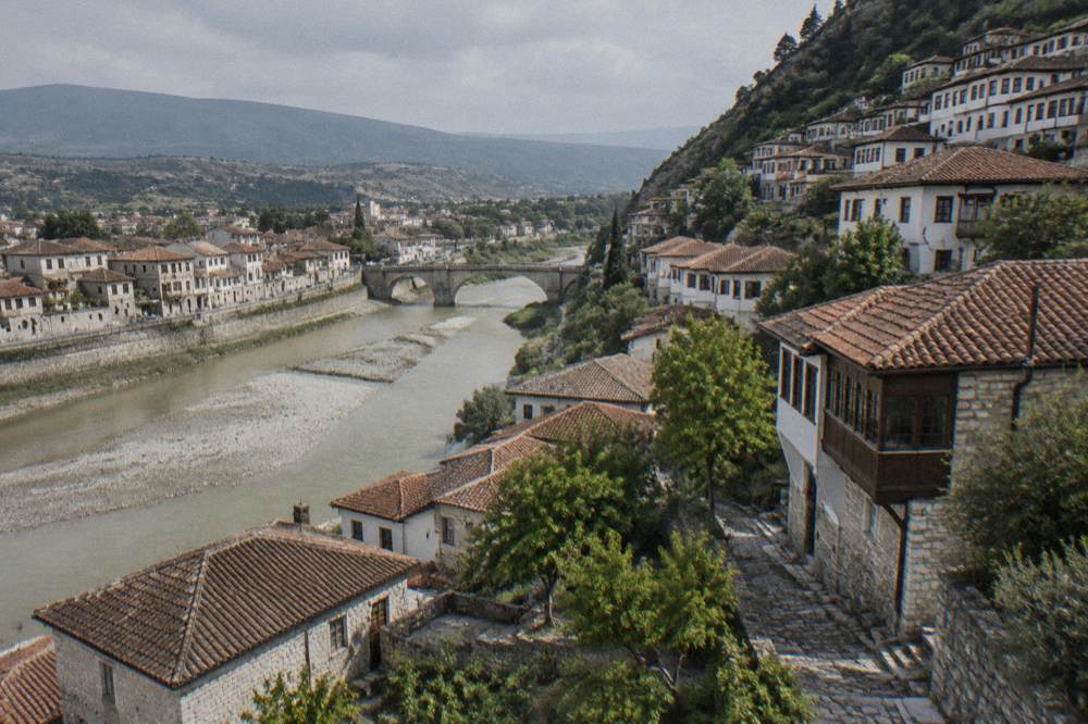 Sunlit stone houses along the Osum River in Berat with the Gorica Bridge in the background