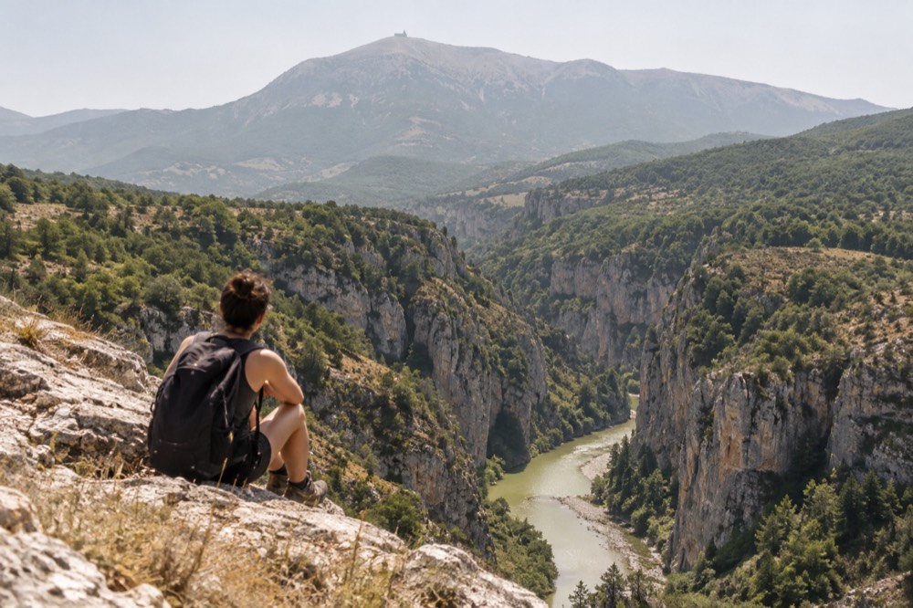 A hiker with a backpack resting at a canyon rim viewpoint above green water