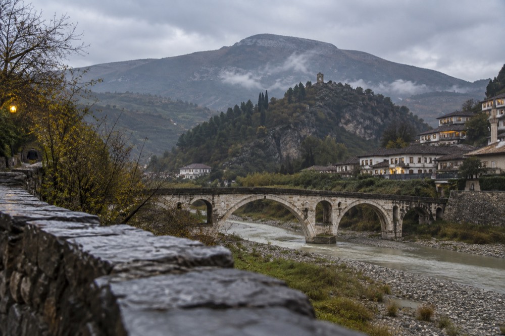 A stone bridge over the Osum River at dusk with hills behind the town