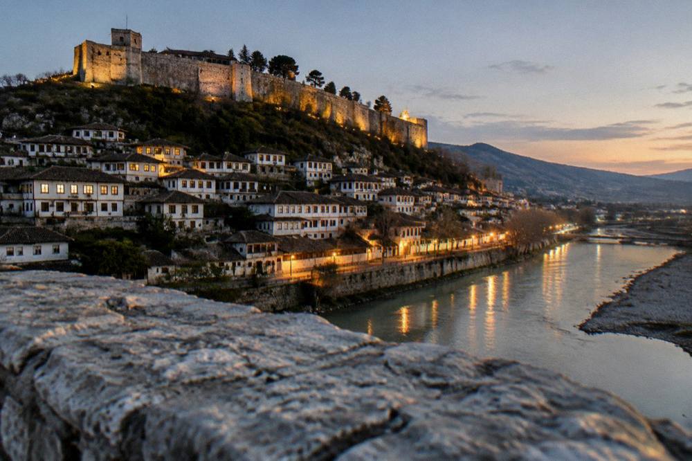 Evening view of a floodlit hillside town with lit windows reflecting on a river
