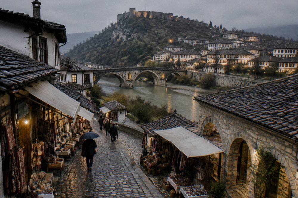 Old stone bridge over the Osum River with lit houses on the far hillside at dusk