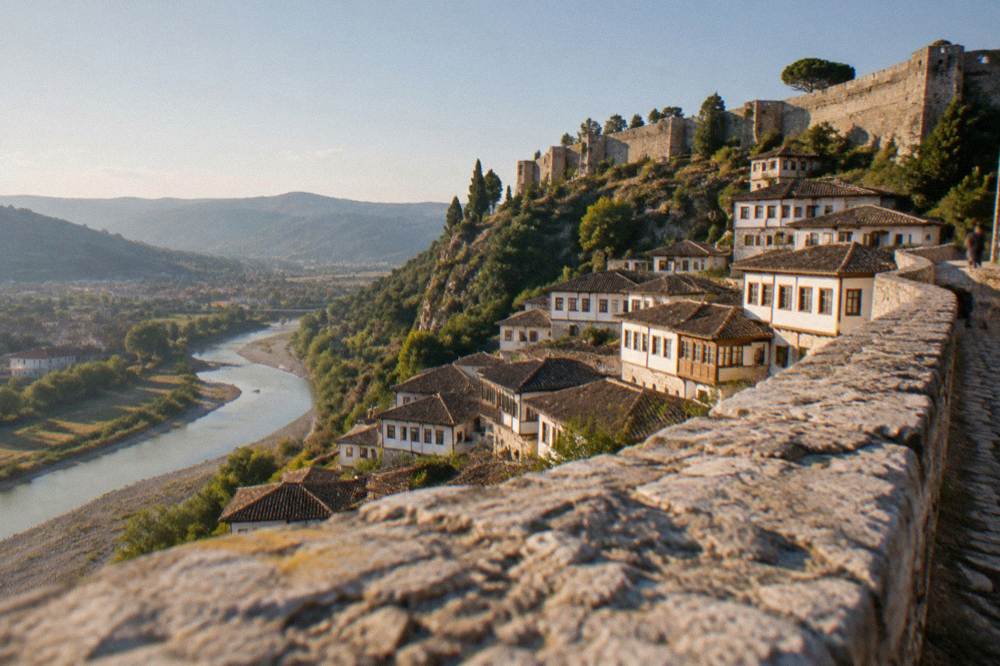 A stone hillside town of white Ottoman houses stacked above a river, with a castle wall on the ridge above.
