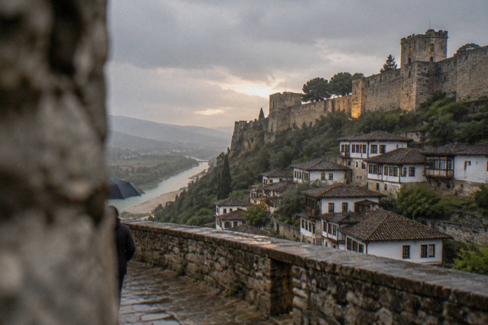 A stone castle wall with towers above white houses on a hillside in the evening light.