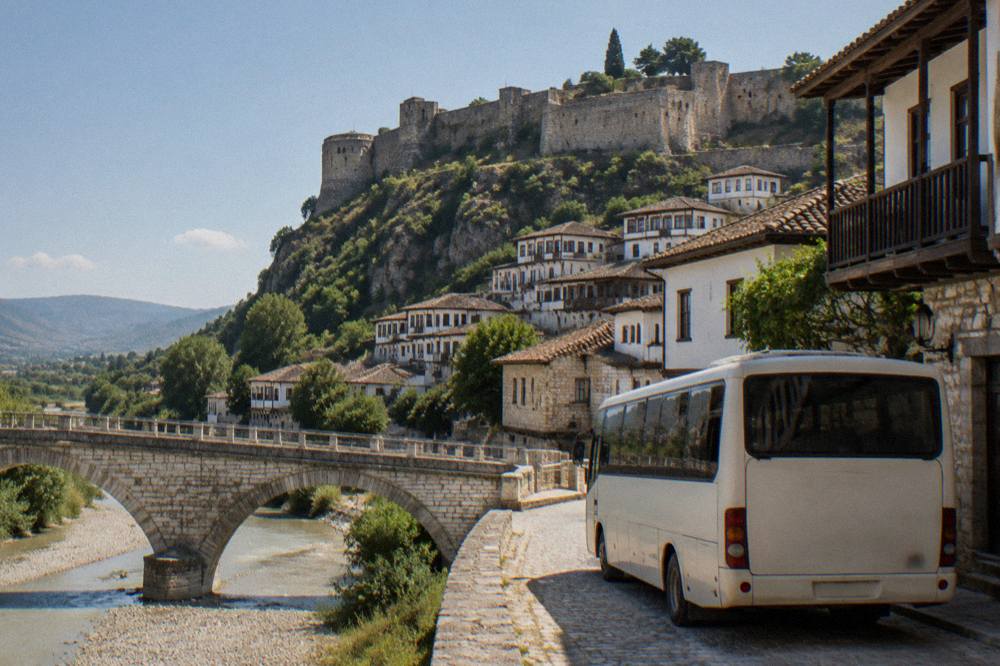 A bus parked on a small town street beside a stone bridge and old houses.