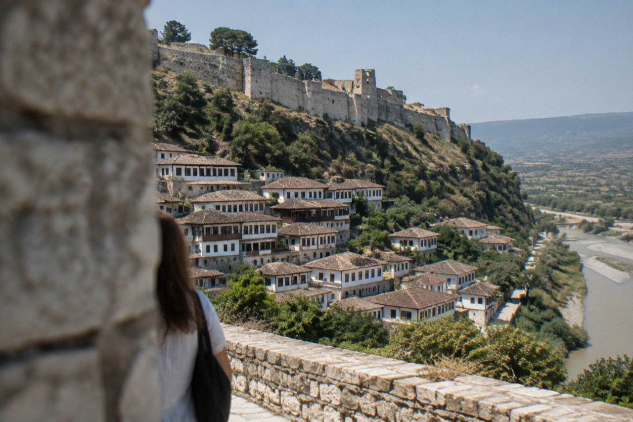 A stone hillside town of white Ottoman houses stacked above a river, with a castle wall on the ridge above.